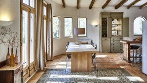 A spacious dining area with a large wooden table, multiple chairs, and a patterned rug. There are large windows with curtains, a built-in shelf with glassware, and a kitchen area visible in the background.