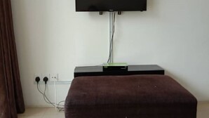 A room with a flat-screen TV mounted on the wall, a power strip, and a brown ottoman on the floor.