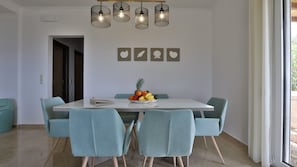 Un coin repas avec une table blanche, quatre chaises bleu clair, un panier de fruits et un lustre à quatre lampes.