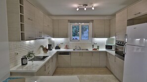 Uma cozinha moderna com armários de madeira clara, bancada branca, máquina de lavar louça embutida, pia dupla, geladeira de aço inoxidável e janela com vista para vegetação.