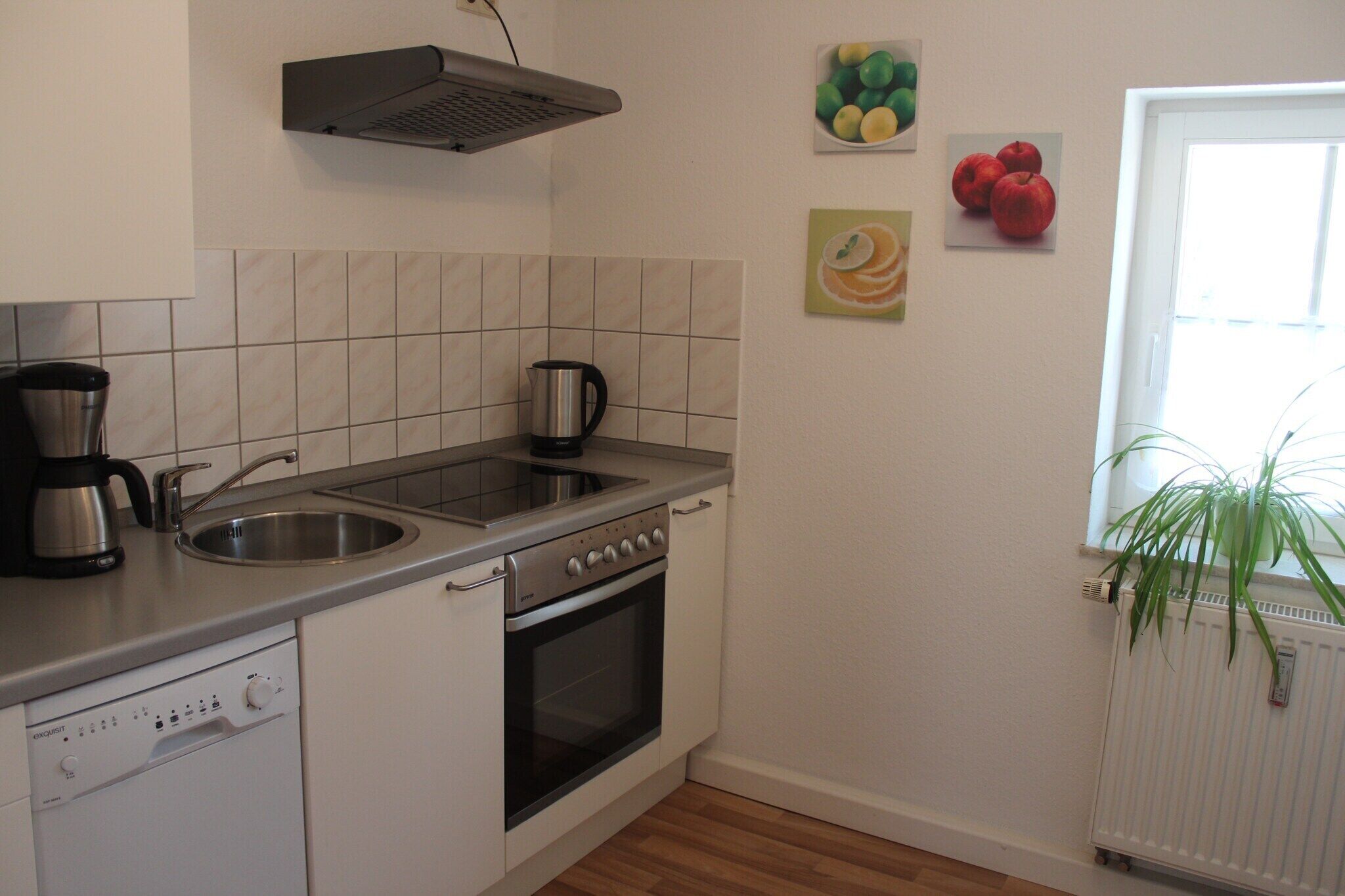 Eine Küche mit Geschirrspüler, Backofen, Spüle und Kaffeemaschine. An den Wänden befinden sich Bilder von Früchten, und in der Nähe des Fensters steht ein Heizkörper.