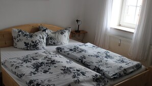 Uma cama de casal com roupa de cama florida, uma mesinha de cabeceira com abajur e uma janela com cortinas.