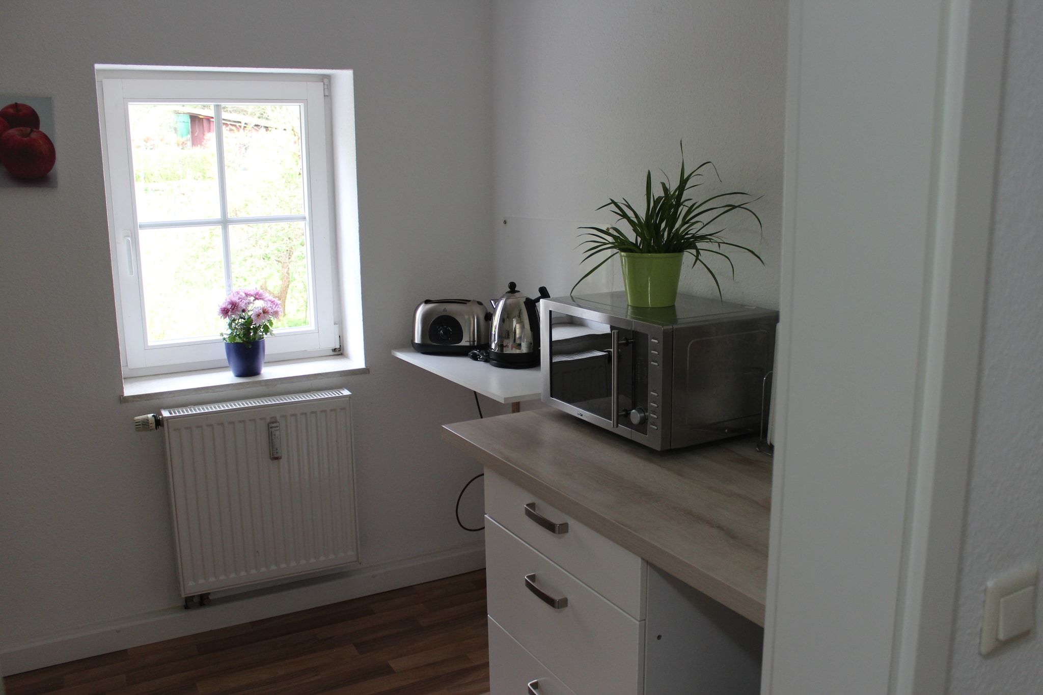 Een keuken met een raam, een radiator, een broodrooster, een waterkoker, een magnetron, een plant in pot en een kast.