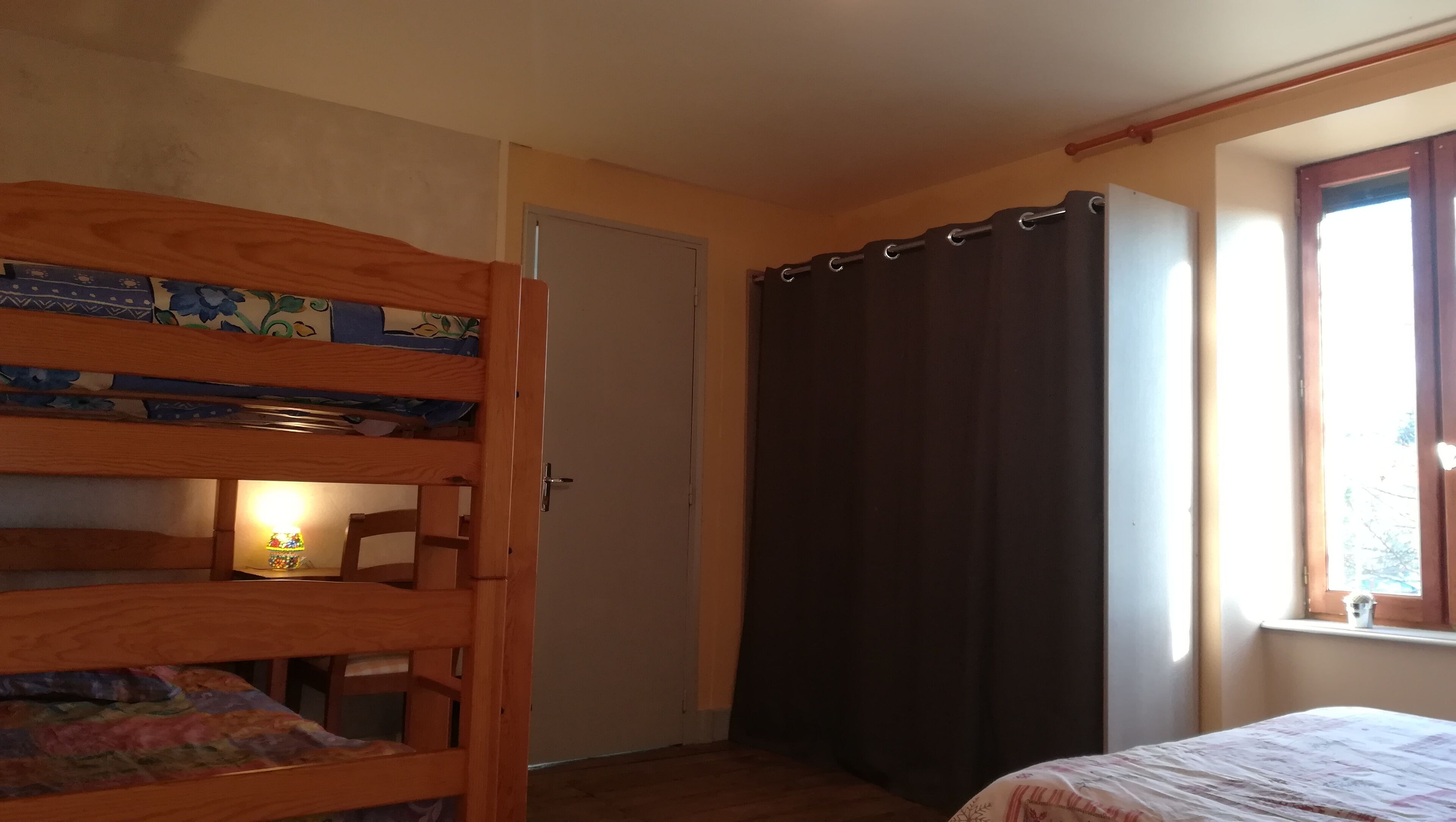 Ein kleiner, gemütlicher Raum mit einem Etagenbett, einer Tür, einem Fenster mit Vorhängen und einem kleinen Tisch mit einer Lampe.