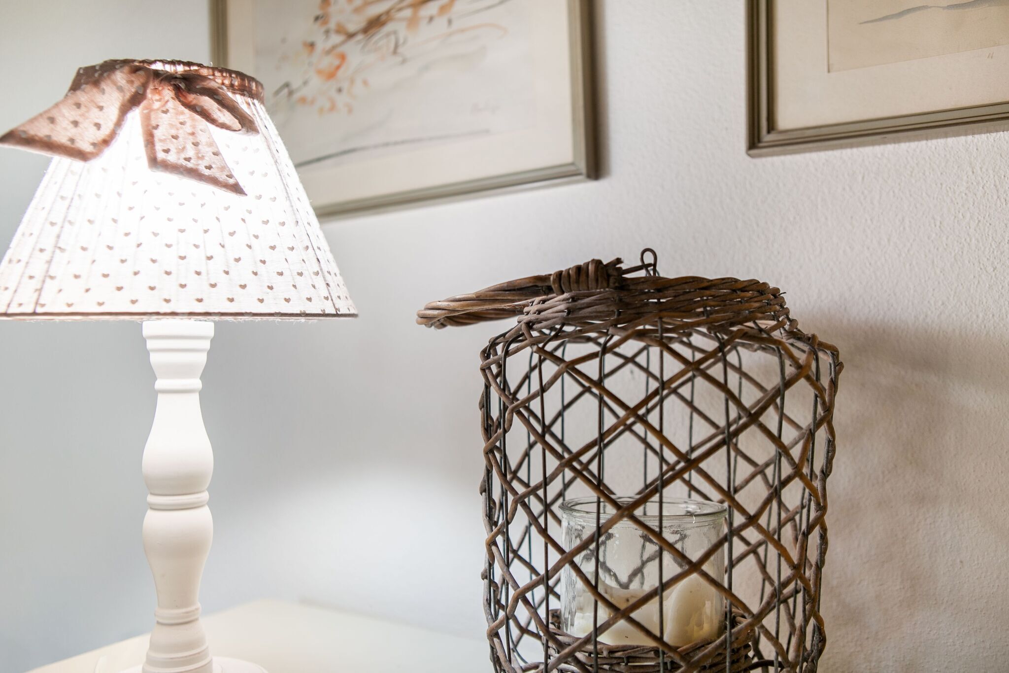 A lamp with a white shade and a decorative bow, a wicker basket with a candle inside, and framed artwork on the wall.