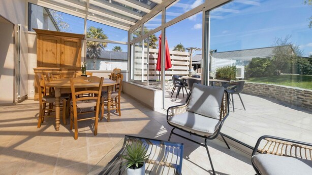 Un patio soleado con mesa y sillas de madera, un espacio cubierto con vidrio que ofrece vistas al jardín trasero, y una zona de estar con una silla acolchada.