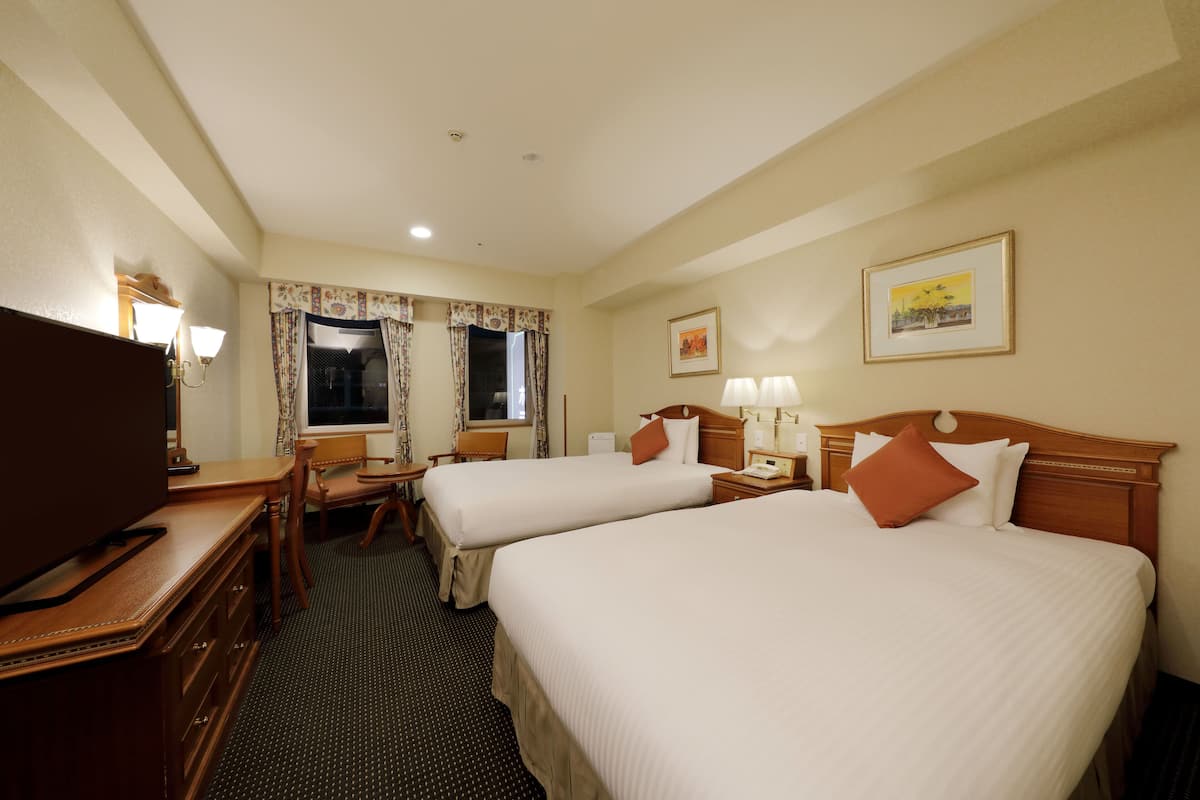 Habitación de hotel con dos camas, un escritorio con televisor, una silla, una lámpara de escritorio y una ventana con cortinas.