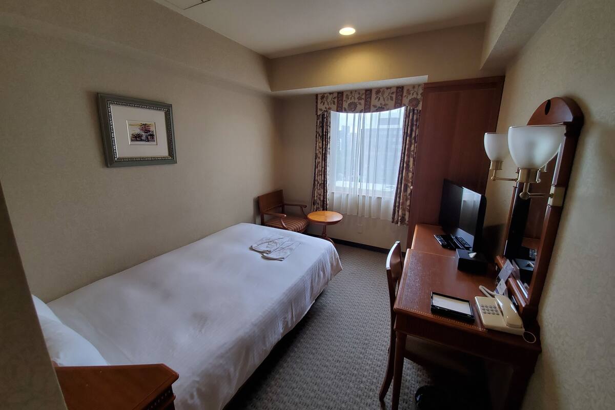 Habitación de hotel con una cama individual, un escritorio con teléfono y lámpara, una silla, una mesita pequeña y un cuadro en la pared.