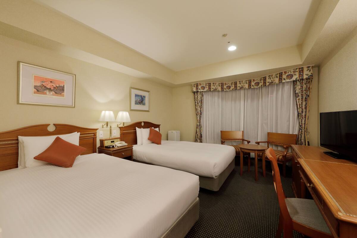 Habitación de hotel con dos camas, un escritorio, una silla, un televisor y dos lámparas.