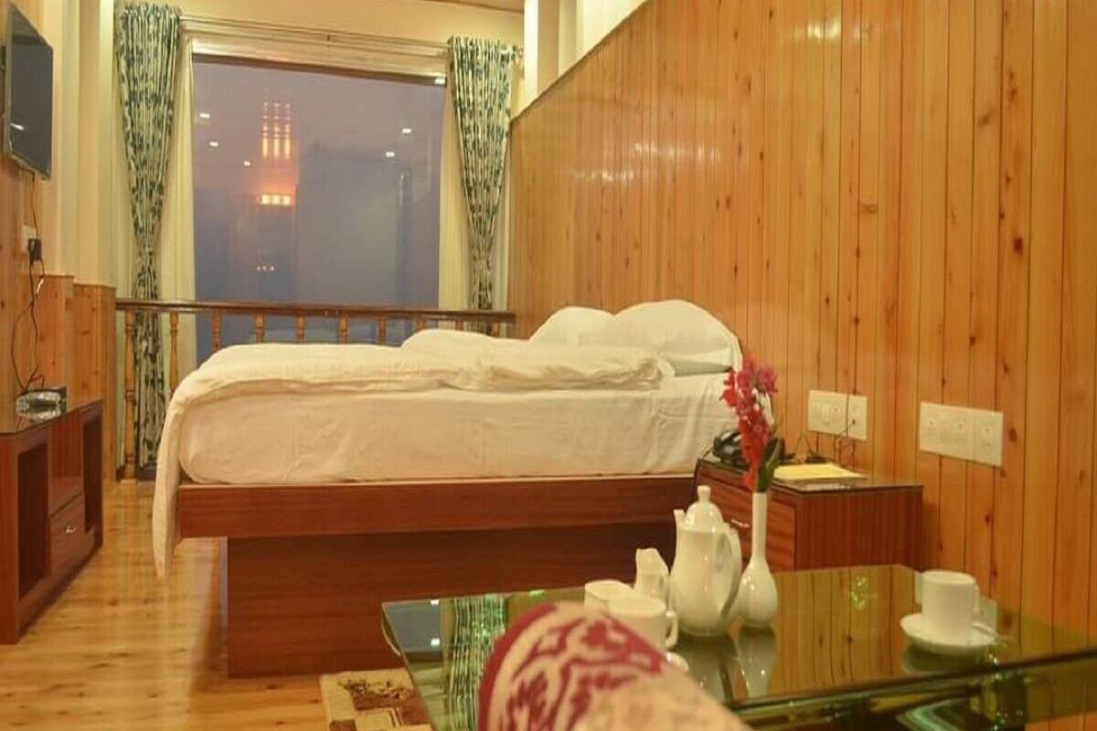 Habitación de hotel con decoración de madera, una cama con ropa de cama blanca, una mesa de vidrio con una tetera y vistas a la ciudad a través de la ventana.