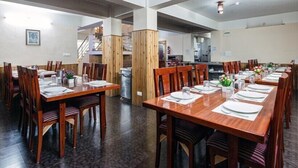 Un comedor amplio con mesas y sillas de madera, una escalera al fondo y una cocina visible a través de una puerta abierta.