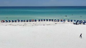 Une plage de sable blanc bordée d’une mer d’un bleu limpide. On y voit plusieurs rangées de parasols bleus et quelques personnes dans l’eau.