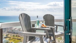 Un balcon avec deux fauteuils Adirondack, une table avec une bouteille et des verres, et une vue sur l’océan et les bâtiments.