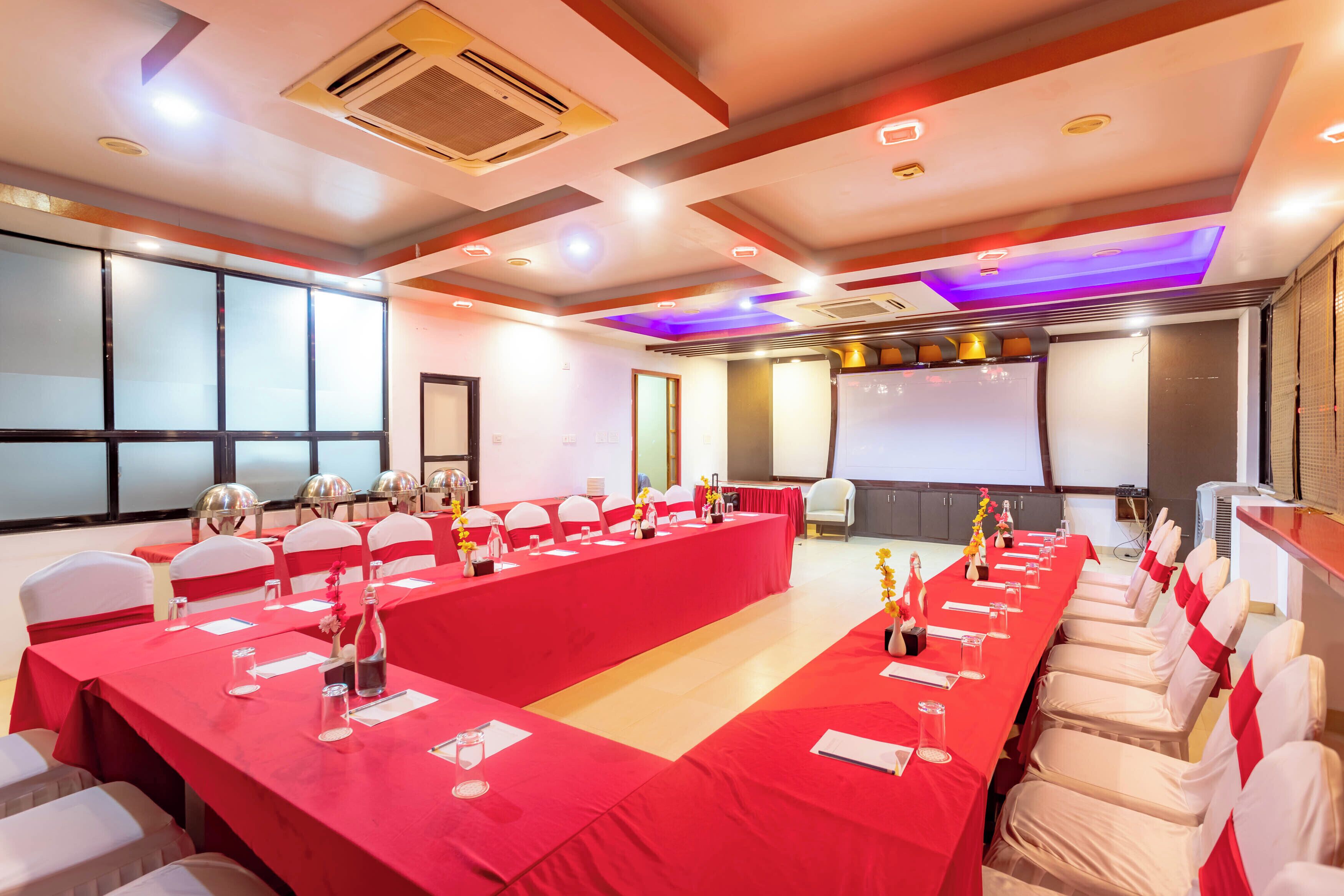 Un salón de conferencias con una larga mesa roja, sillas blancas y una pantalla de proyector.