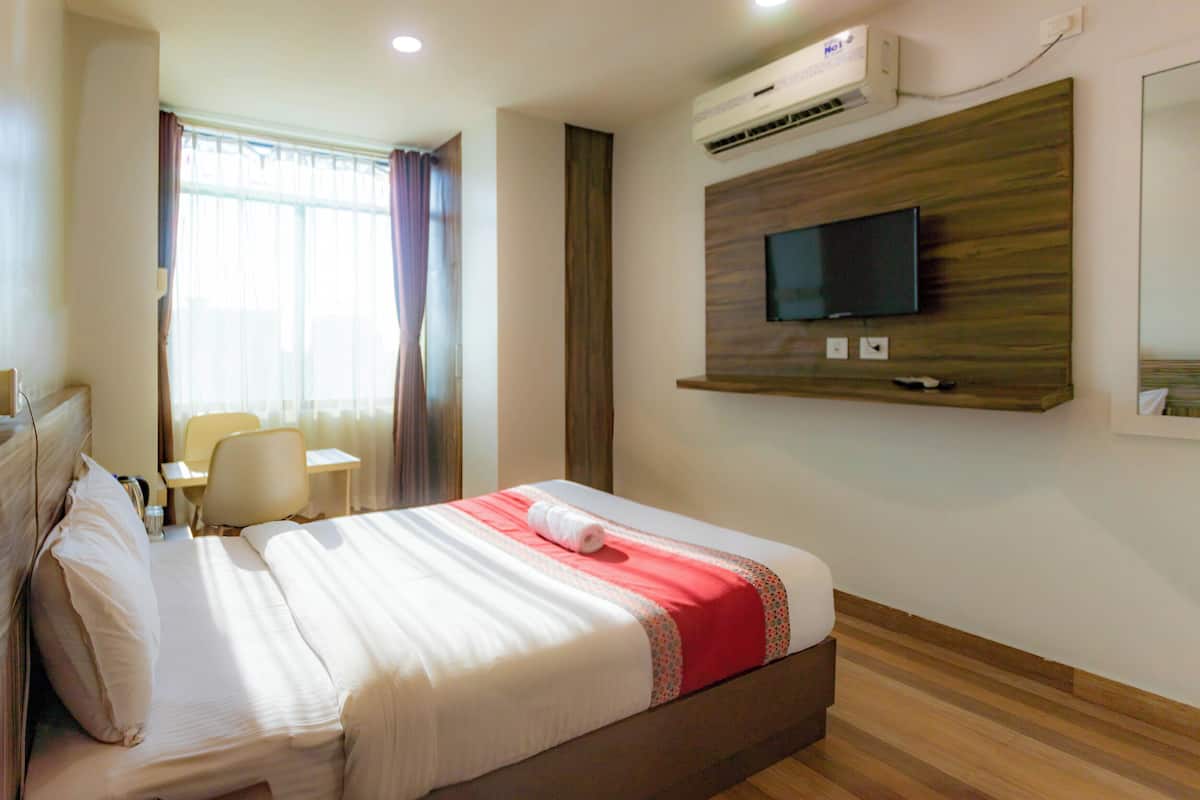 A hotel room with a bed, a desk, a chair, a television mounted on the wall, and an air conditioning unit.