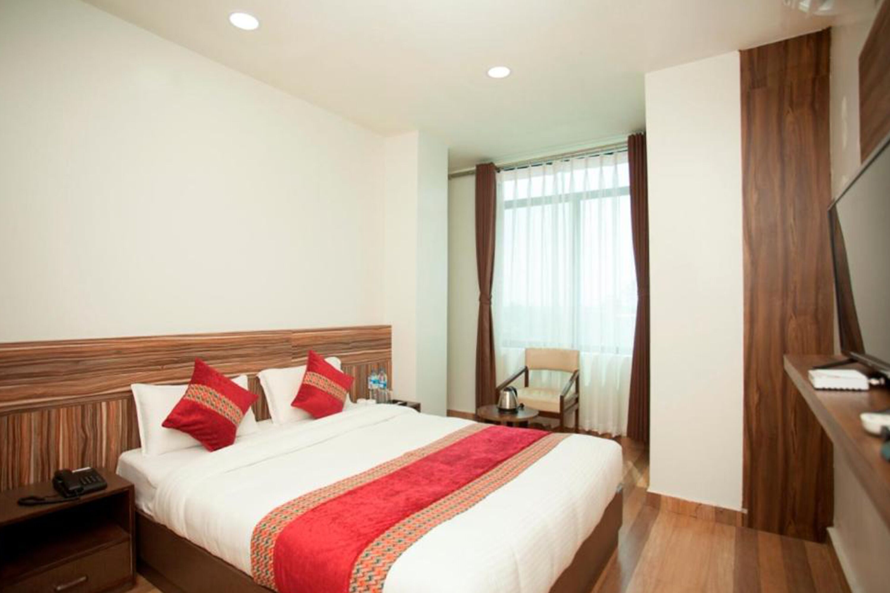 A hotel room with a large bed, wooden headboard, a TV on a stand, and a chair near the window.