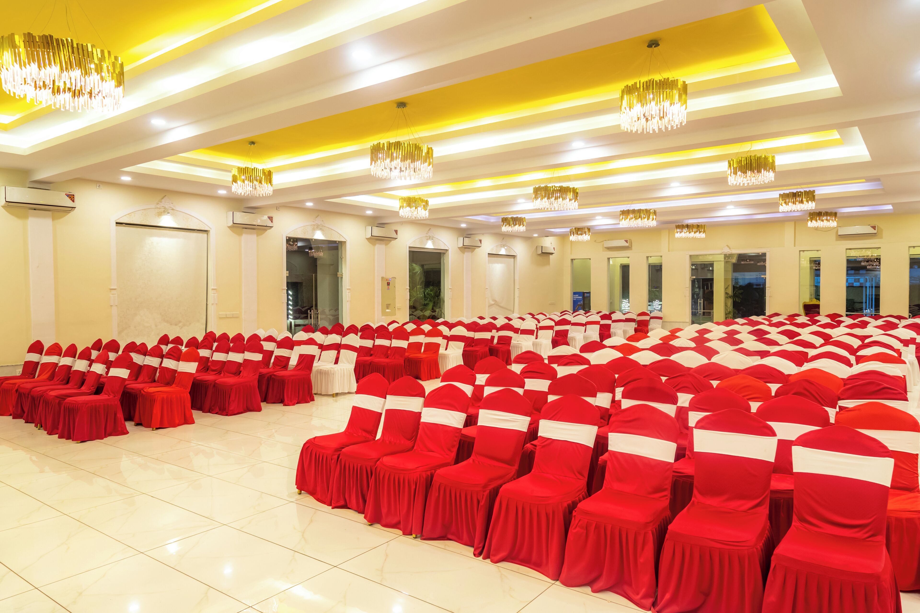 Un gran salón con hileras de sillas rojas y lazos blancos, unidades de aire acondicionado y candelabros.