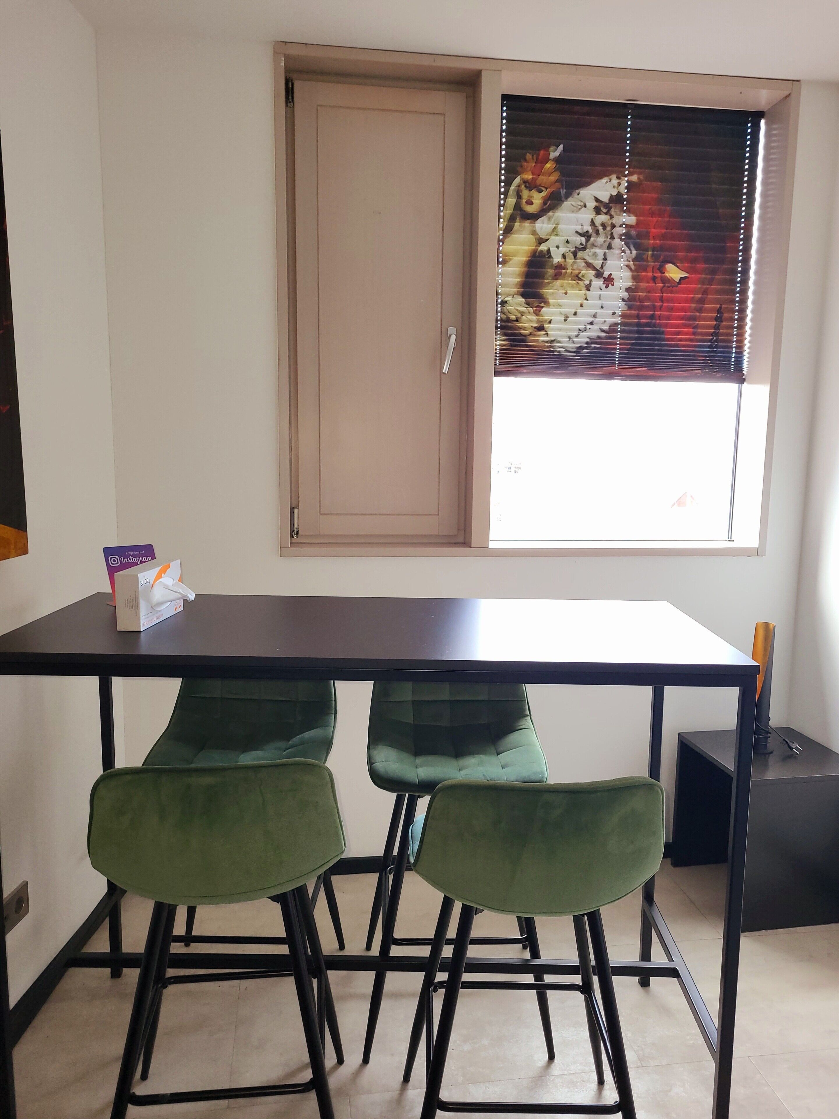 Ein Zimmer mit Tisch, zwei Stühlen, einer Tür und einem Fenster mit Jalousien.