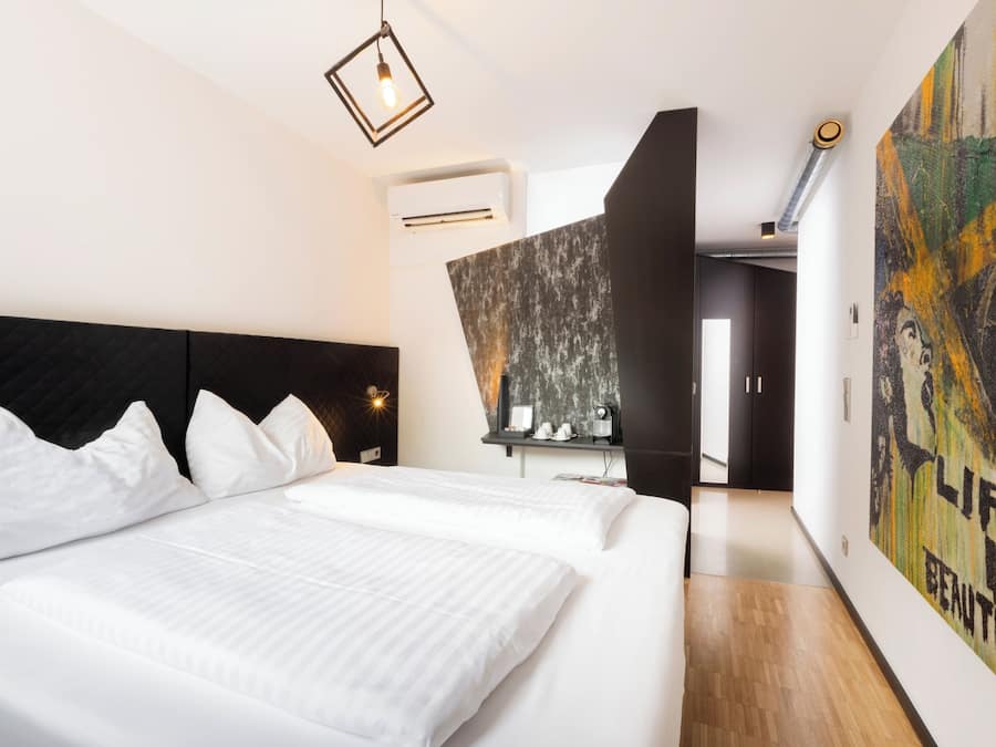 Et moderne hotelværelse med en stor seng, et sort sengegavl, en vægmonteret aircondition og et stort abstrakt maleri.