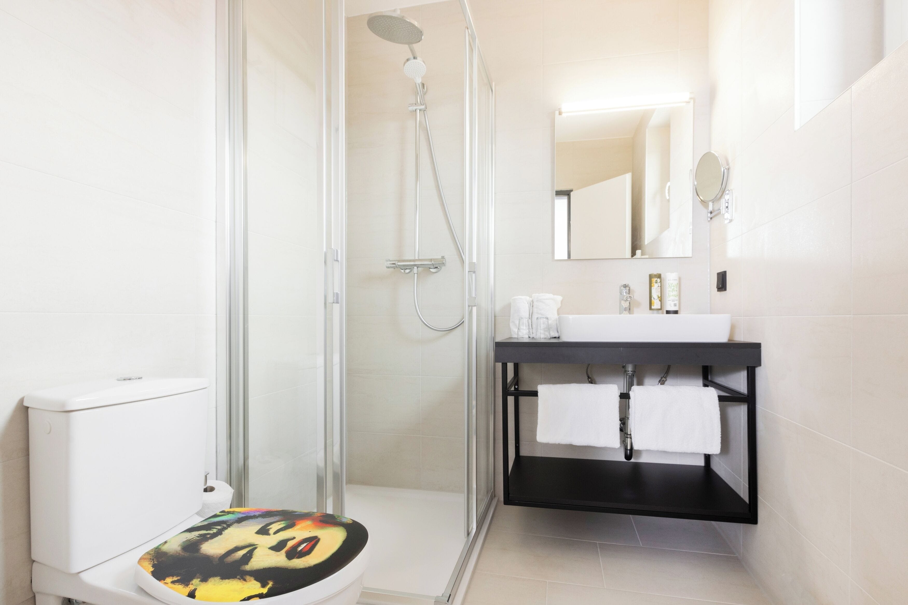 Une salle de bain moderne avec une douche à parois de verre, des toilettes blanches dotées d’un abattant coloré, un lavabo noir avec un miroir au-dessus, et une serviette blanche accrochée au mur.