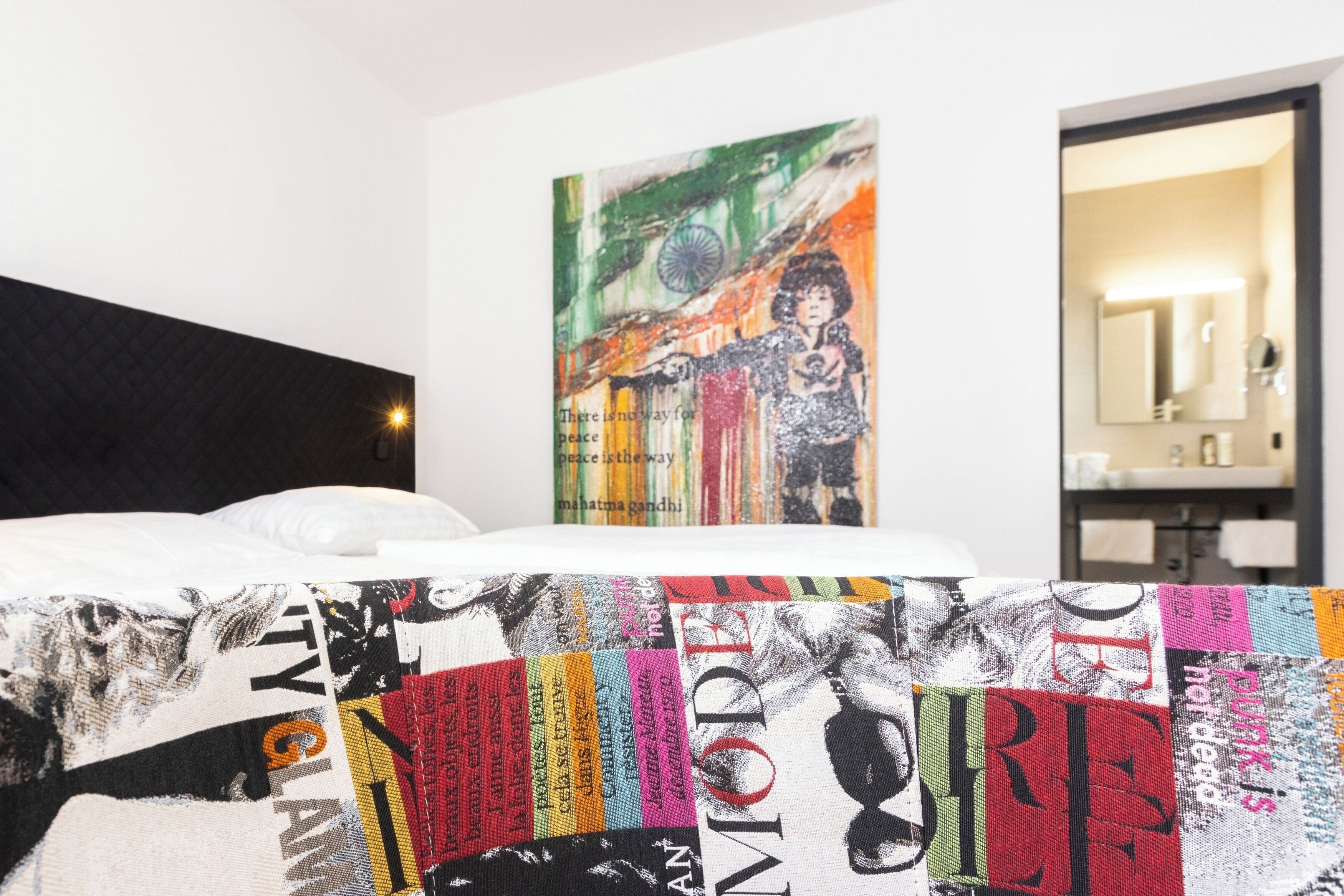 A hotel room with a bed, a colorful artwork on the wall, and a bathroom with a mirror and sink.