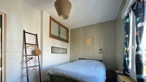 Een slaapkamer met een bed, een houten trapje, een hanglamp, een raam met gordijnen en een radiator.