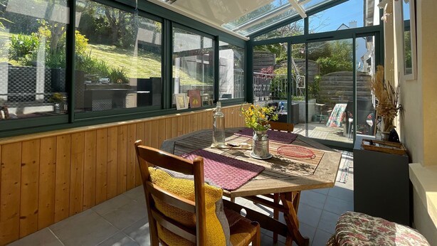 Een orangerie met een houten tafel en stoelen, een bloevas, en uitzicht op een tuin.