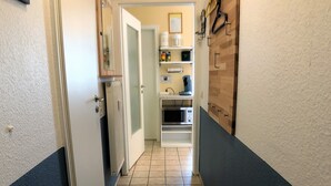 Un pasillo estrecho con suelo de baldosas, paredes blancas y una pared azul decorativa. Hay una puerta a la izquierda, una estantería con varios objetos a la derecha, y un microondas en el suelo.