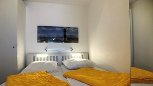 Una cama individual pequeña y bien hecha con sábanas blancas y una manta amarilla, cabecera blanca y un cuadro colgado en la pared con un faro.