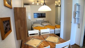 Una habitación pequeña con una mesa de madera, dos sillas blancas, una cama con cabecera blanca, un espejo y una lámpara en el techo.