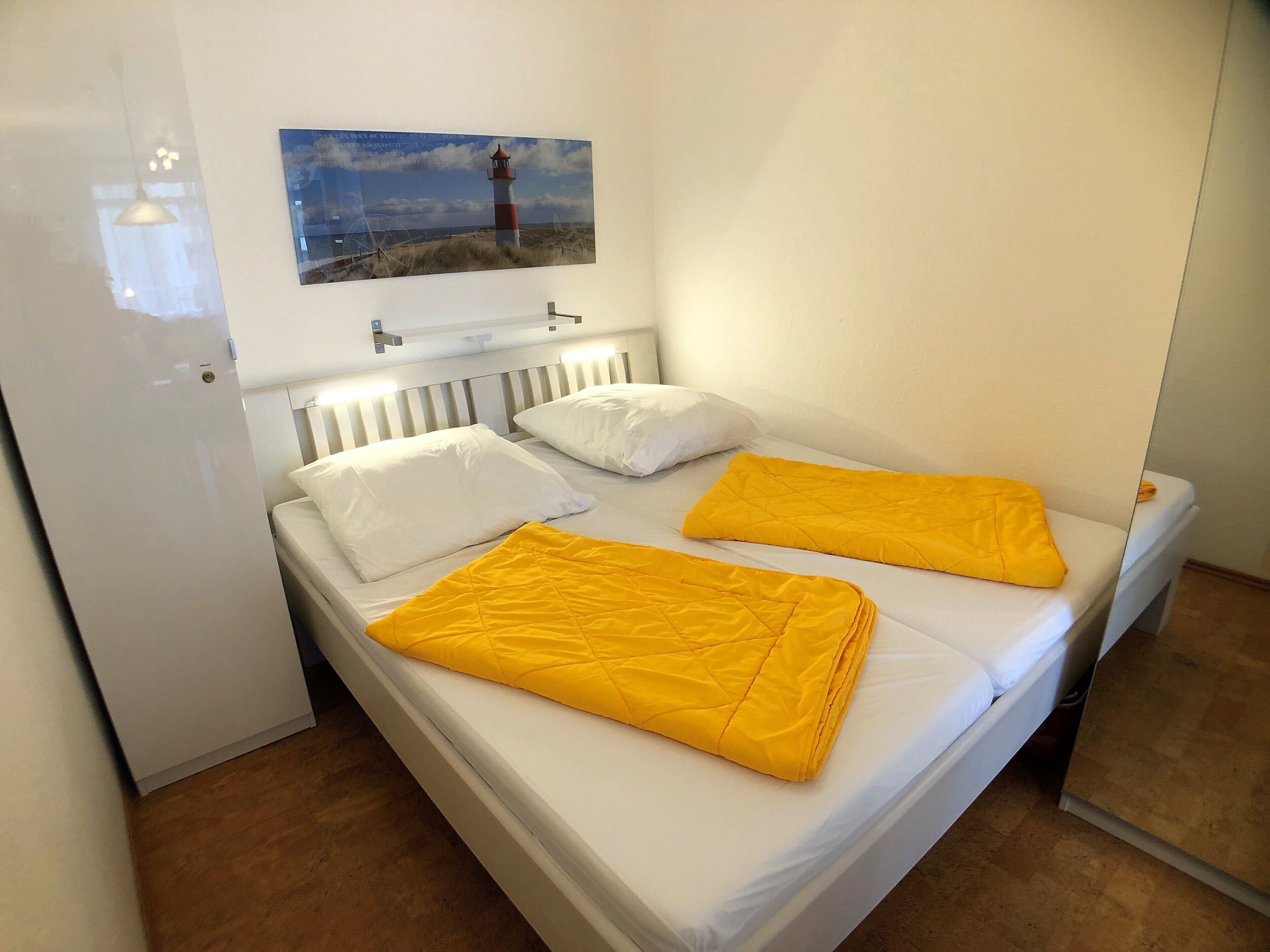 Un lit double avec une literie blanche et deux couvertures jaunes, une tête de lit blanche et un tableau représentant un phare accroché au mur.