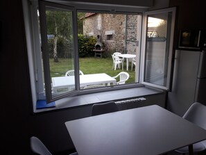 A room with a large window overlooking a garden with a stone building, a barbecue, and outdoor furniture.