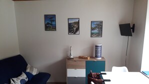 Ett rum med en blå soffa, ett träskåp med en vit och turkosfärgad låda, ett vitt bord och en lampa. På väggen hänger tre inramade tavlor.