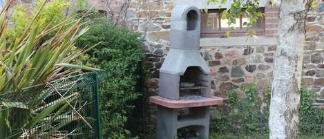 Une structure de four extérieur en pierre, avec une base en briques, entourée de verdure et d’un mur de pierres.