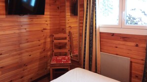 Une pièce de style cabane en bois, avec un lit, un téléviseur fixé au mur, une chaise avec un coussin rouge et une fenêtre avec des rideaux.