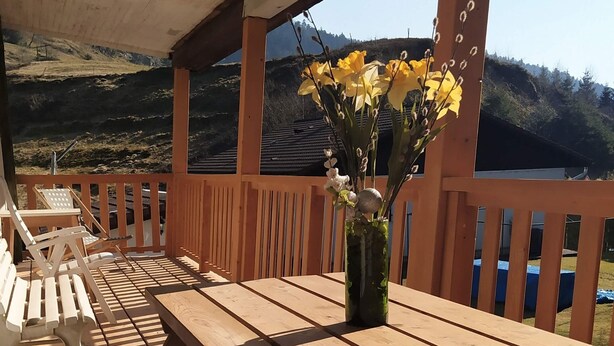 Une terrasse en bois avec une table et des bancs, un vase rempli de fleurs jaunes et une vue sur un paysage vallonné.