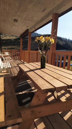 Een houten terras met een tafel en bankjes, een vaas met gele bloemen, en uitzicht op een glooiend landschap.