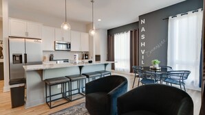Une cuisine moderne avec des armoires blanches, un réfrigérateur en acier inoxydable et un four à micro-ondes. On y trouve un coin bar avec des tabourets et une table à manger avec des chaises. La pièce est dotée d’une grande fenêtre avec des rideaux et d’un mur sur lequel est inscrit le mot « NASHVILLE ».