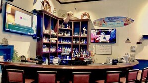 Zona de bar amb un taulell de bar de fusta, diverses estanteries plenes de botelles, una pantalla de televisió i una decoració amb una taula de surf.