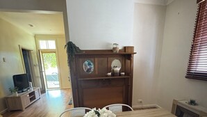 A dining area with a wooden table, chairs, and a vase of white flowers. A television is visible in the background.