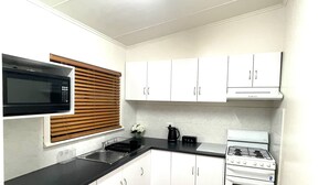 A compact kitchen with white cabinets, a black countertop, a microwave, a stove, and a window with blinds.