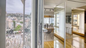 A modern apartment with large windows offering a city view, a wooden floor, and a dining area with a table and chairs. - Sunny Luxury Loft, Stunning Acropolis View, Jacuzzi (Athina)