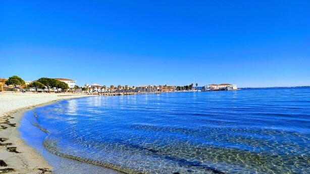 A beach with clear blue water, a sandy shore, and buildings in the background.
