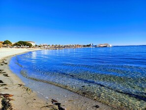 En strand med kristallklart blått vatten, en sandstrand och byggnader i bakgrunden.