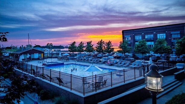 A rooftop pool area with lounge chairs and umbrellas, surrounded by a fence, under a sunset sky.