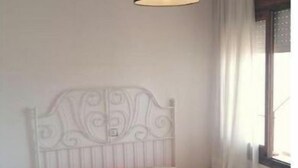 Un dormitorio con cabecera de metal blanco, cama con colcha a rayas, mesita de noche, lámpara colgante y ventana con cortinas.