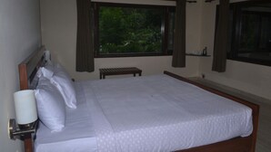 A neatly made bed with white linens and pillows, a bedside table with a lamp, and a window with curtains. - Corbett Comfort Lodge (Ramnagar)
