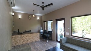 A room with a reception desk, two sofas, a coffee table, a ceiling fan, and a view of greenery outside. - Corbett Comfort Lodge (Ramnagar)