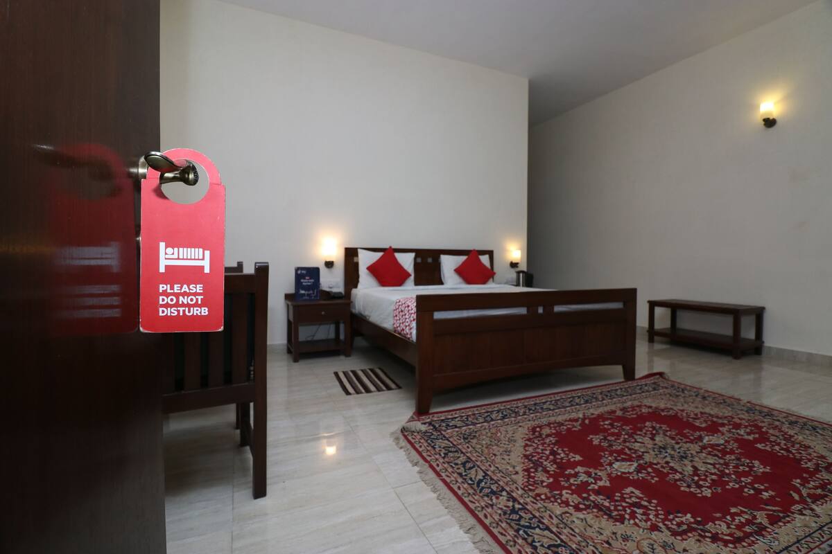 A hotel room with a bed, bedside tables, a nightstand, a red rug, and a 'Please do not disturb' sign.
