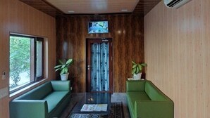 A room with a wooden ceiling, a ceiling fan, a television mounted above a door, and two green sofas. - Corbett Comfort Lodge (Ramnagar)